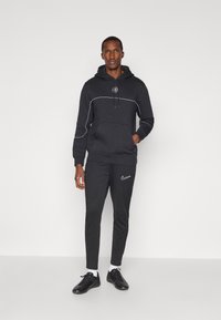 Melns kapuce ar baltām akcentu līnijām un logo, pāris ar atbilstošām melnām biksēm, kurām ir mazs Nike logo. Modelis valkā melnus sporta apavus.