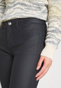 Jeans ajustés bleu marine avec une texture lisse, présentant des coutures détaillées, des poches avant et un bouton en métal à la taille.