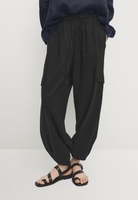 Mustat kevyet housut, joissa löysä istuvuus, joustava vyötärönauha, sivutaskut ja kerätyt lahkeensuut. Yhdistetty mustien tasapohjaisten sandaalien kanssa.