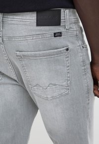 Lysegrå denimjeans på en person, som viser bak høyre lomme med knapp og to svarte merkeetiketter over.