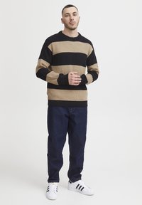 Pleteni pulover s horizontalnimi črtami v črni in bež barvi, okroglim ovratnikom in rebrasto teksturo. Usklajen z temnimi denim hlačami in belimi čevlji.