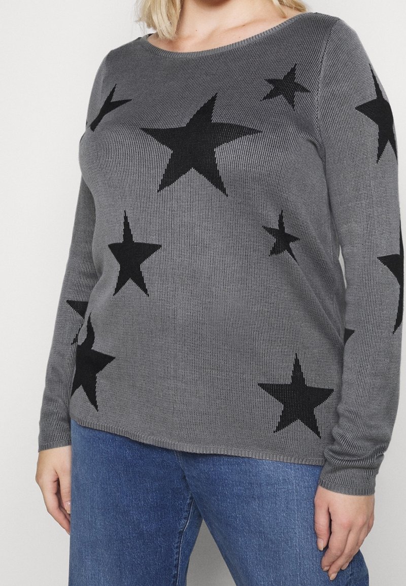 Pull en maille gris avec des motifs d'étoiles noires porté avec un jean bleu, montrant le torse et les bras, sans visage ni détails de l'arrière-plan visibles.