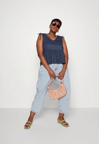 Top peplum azul-marinho com alças para amarrar, jeans largos azul claros, sandálias verdes e uma mala bege. Óculos de sol usados. Fundo neutro.