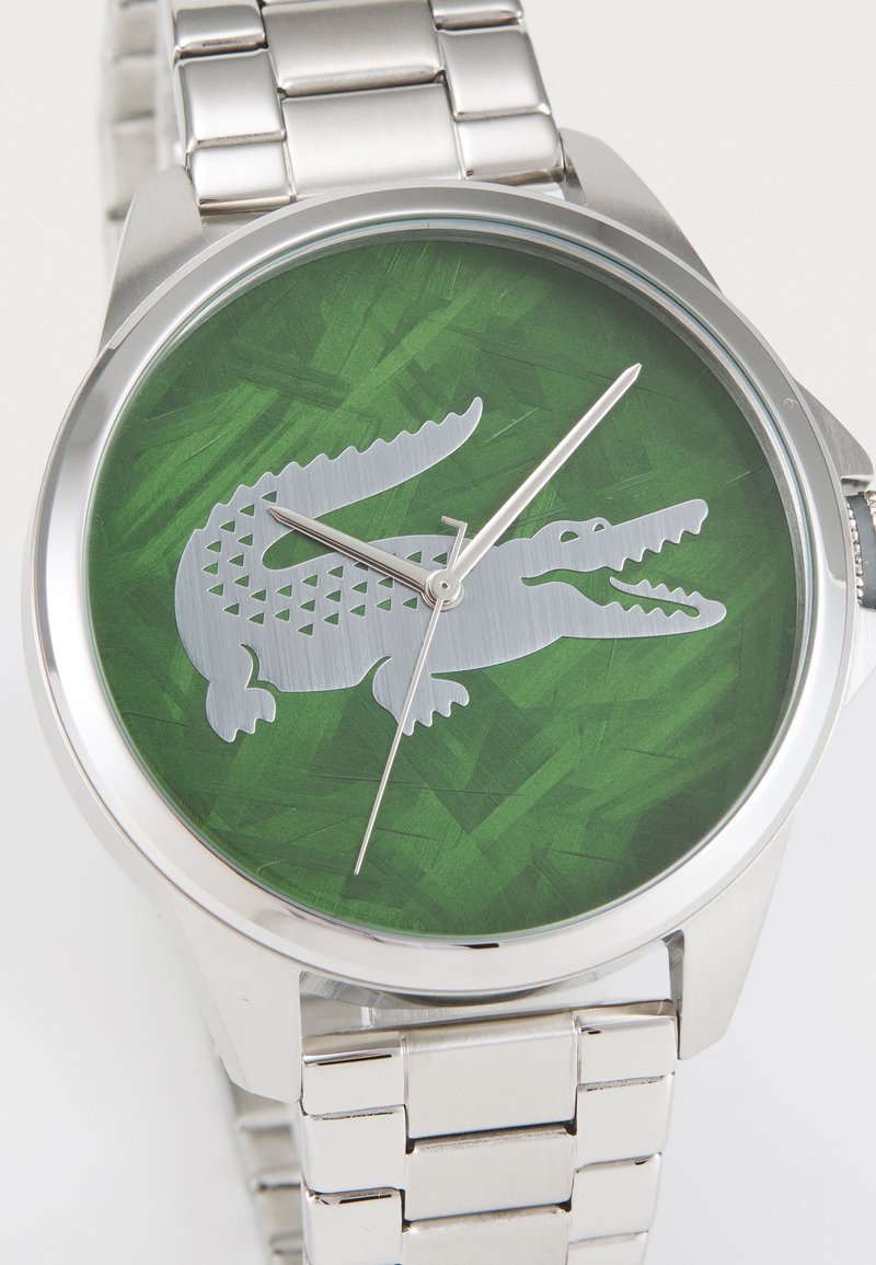Reloj de acero inoxidable plateado con un dial texturizado verde que presenta un gráfico de cocodrilo plateado y agujas plateadas delgadas. Correa de malla.
