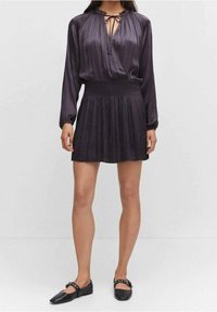 Lila plissiertes Kleid mit einer Bluse mit Bindeband, langen Ärmeln und smockierter Taille. Schwarze flache Schuhe mit dekorativen Riemen. Weiche Textur.
