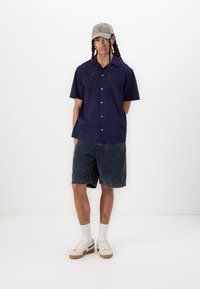 Camisa de manga corta en color marino con detalles bordados, combinada con unos pantalones cortos de mezclilla. La modelo lleva una gorra beige y zapatillas blancas con suelas marrones.