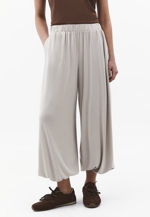 Stehende Person trägt lockere, hellgraue, weit geschnittene Hosen mit elastischem Bund und braune Sneakers, Hände teilweise in den Taschen, vor weißem Hintergrund.