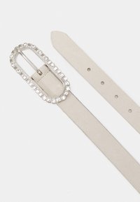 Ceinture en cuir beige avec une boucle ovale en métal argenté ornée de strass transparents et plusieurs trous de réglage le long de la sangle.
