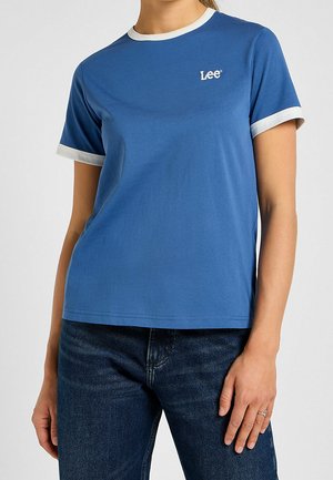Kobieta ubrana w niebieski t-shirt Lee z białym obszyciem na rękawach i kołnierzu, w połączeniu z ciemnoniebieskimi jeansami i pierścionkiem na lewej ręce.