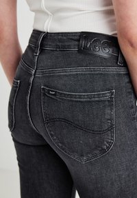 Black denim jeans featuring a high-waisted design, back patch pocket with a logo, and subtle stitching details on the pockets.