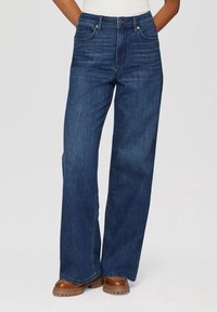 Persona vistiendo jeans de tiro alto de pierna ancha de color azul oscuro y zapatos de suela gruesa marrones, con las manos metidas en los bolsillos.
