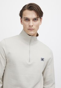 Beige zip-up sweatshirt with a high collar, made of soft fabric. Features a small, dark logo on the left chest area.
