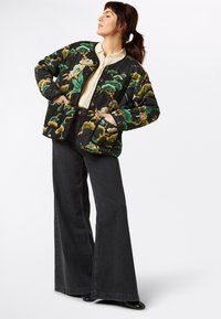 Gequilted zwarte jas met een bloemig patroon in groen en geel, voorzien van contrasterende bies, twee zakken en een relaxte pasvorm. Gecombineerd met flared jeans.
