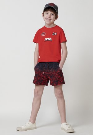 Maglietta rossa a maniche corte con grafica stampata e numero 24, abbinata a pantaloni corti a righe nere e rosse e sneakers bianche. Cappellino grigio.