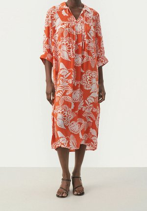 Femme portant une robe orange ample, longueur genou, avec un grand imprimé floral blanc et des sandales noires à brides, debout sur un sol clair.