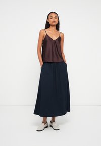 Kobieta w bezrękawnikowej, ciemnoniebieskiej bluzce, długiej granatowej spódnicy z kieszeniami oraz jasnobeżowych butach wsuwanych, stoi przy gładkiej białej ścianie.