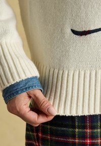 Krēmveida adīts džemperis ar ribotām piedurknēm un apakšmalu; zem tā ir zila denim krekla slānis ar kareījo rakstu apakšā.