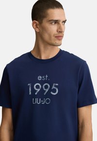 T-shirt in cotone blu navy con maniche corte, con una stampa grigia invecchiata di "est. 1995 LIU•JO" centrata sul davanti.