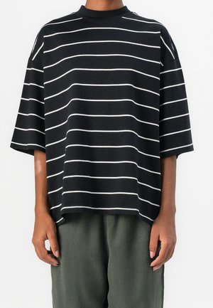 Persoon draagt een loszittend zwart shirt met witte horizontale strepen en donkergroene broek, staat met ontspannen armen langs het lichaam.