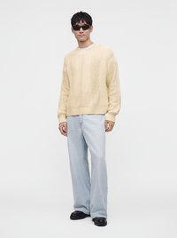 Jeune homme portant des lunettes de soleil noires, un pull en maille torsadée crème, un jean large bleu clair et des chaussures noires, debout devant un fond uni.