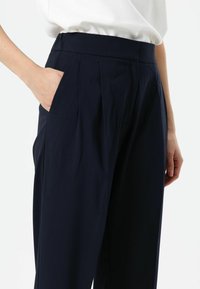 Marineblauwe plissébroek met een gladde stoftextuur, voorzien van zijzakken en een hoge tailleband, afgebeeld met een hand in de zak.