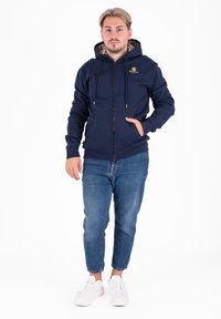 Felpa con zip di colore navy, con interno a quadri, cordini e tasche frontali, abbinata a jeans azzurri chiari e sneakers bianche.