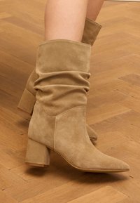 Tan suède enkellaarsjes met een slouchy ontwerp, puntige neus en blokhak, tegen een houten vloer.