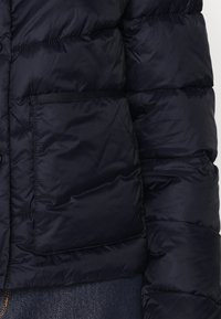 Černá péřová bunda se širokým čtvercovým kapsou na rukávu, viditelné švy, nošená přes tmavě modrou džínovou látku.