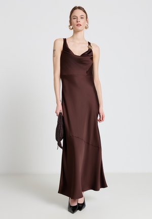 Femme portant une robe longue en satin marron foncé sans manches avec de larges bretelles, tenant une pochette tissée foncée et portant des chaussures noires pointues.