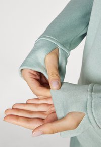 Sudadera verde claro con agujeros para los pulgares en las mangas, hecha de una tela suave y texturizada. Vista en primer plano de manos ajustando el puño.