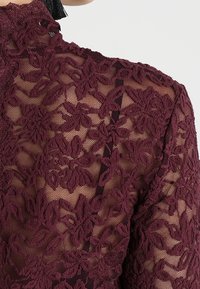 Burgundy spetsblus med blommönster och genomskinlig textur. Har hög hals och intrikata detaljer längs axlarna och ärmarna.
