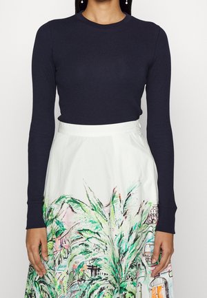 Kvinde iført en marineblå top med lange ærmer og hvid nederdel med farverige grønne og lyserøde tropiske blade og bymønstre.