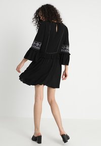 Vestido negro con mangas acampanadas, con detalles bordados en color en los puños y un dobladillo con volantes. Diseño de abertura en forma de ojo de cerradura en la parte posterior.