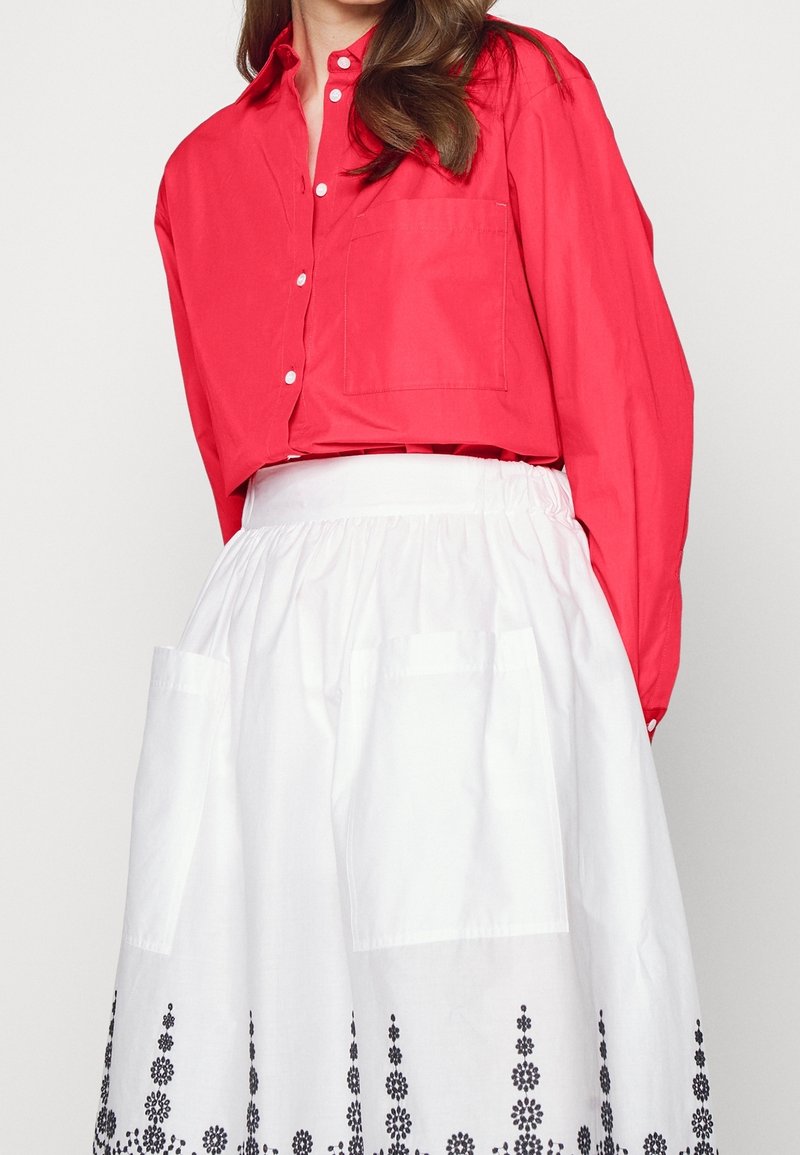 La femme porte une chemise boutonnée rouge vif rentrée dans une jupe blanche avec de grandes poches et une broderie florale noire le long de l'ourlet.