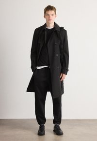 Schwarzer doppelter Trenchcoat mit großen Knöpfen über einem schwarzen Sweatshirt und einem weißen T-Shirt, kombiniert mit schwarzen Jogginghosen und Sneakern.