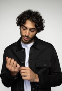 Zwarte button-up jas gemaakt van gestructureerde stof, met een borstzak en een kraag. Daaronder is een effen wit shirt zichtbaar.