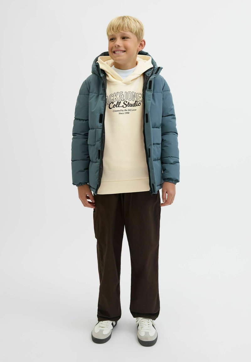 Nasmejan deček v turkizni pufi jakni, kremasti kapuci z besedilom, temnih hlačah in belih supergah, stoji pred enostavno svetlo ozadjem.