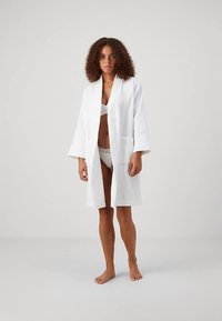 Balts peldmētelis no viegla auduma ar garām piedurknēm, kabatām un brīvu piegriezumu, valkāts pār atbilstošu krūštura un apakšveļas komplektu.