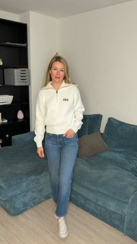 Žena v bílém svetru GAP a modrých džínách prochází kolem tyrkysové manšestrové pohovky v obývacím pokoji se světlou dřevěnou podlahou a černou policovou sestavou.