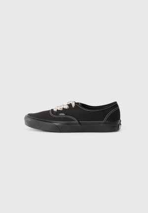 Musta kankaansneaker valkoisilla ompeleilla, pyöreällä kärjellä ja teksturoidulla kumipohjalla. Sisältää mustat nauhat ja logon sivussa.