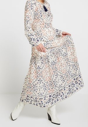 Femme portant une longue robe à motifs floraux avec des pampilles, soulevant légèrement la jupe, assortie à des bottines blanches à la cheville.
