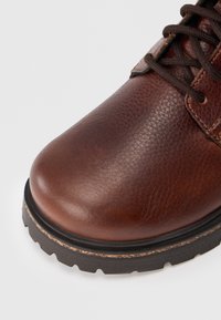 Primer plano de una bota de cuero marrón con cordones, con superficie texturizada y suela de goma negra gruesa sobre fondo blanco.