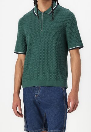 Mann trägt eine strukturierte grüne kurzärmelige Poloshirt mit Reißverschluss und Kragen, kombiniert mit blauen Jeans-Shorts.