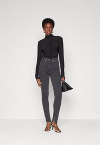 Black long-sleeve bodysuit with gathered detail and high neckline, paired with high-waisted dark gray jeans featuring side slits and black heels.