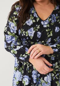 Zwart bloemenjurk met blauwe en groene bloemen; smock-bodice, diepe V-hals en gathering lange mouwen; model draagt een ring.