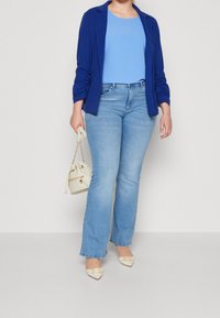 Blazer leggero blu sopra un top azzurro chiaro, abbinato a jeans flare in denim chiaro e una borsa color crema. Ballerine crema a punta con dettagli.