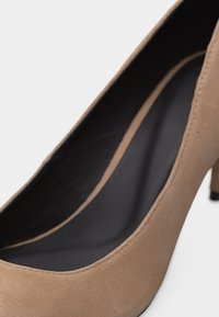 Κοντινή λήψη ενός μπεζ σουέτ γυναικείου παπουτσιού με ψηλό τακούνι, εστιάζοντας στην μαύρη εσωτερική σόλα και στη λεπτομέρεια της καμπυλωτής ραφής.
