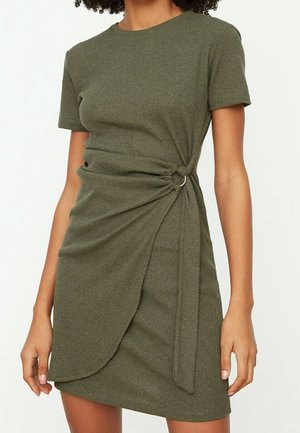 Kvinde iført olivengrøn, ribstrikket kjole med korte ærmer og sammenrynket omslagsdetalje samlet ved taljen med en sølvring.
