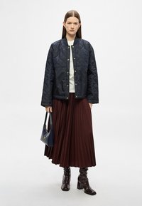 Tengerészkék steppelt bomberdzseki arany patentgombokkal, fehér felső fölött viselve, bordó pliszírozott szoknyával és sötét bokacsizmával párosítva.