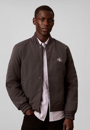 Νέος άνδρας που φορά σκούρο γκρι bomber μπουφάν Calvin Klein πάνω από ένα ανοιχτό ριγέ πουκάμισο με κουμπιά, με τα χέρια στις τσέπες.